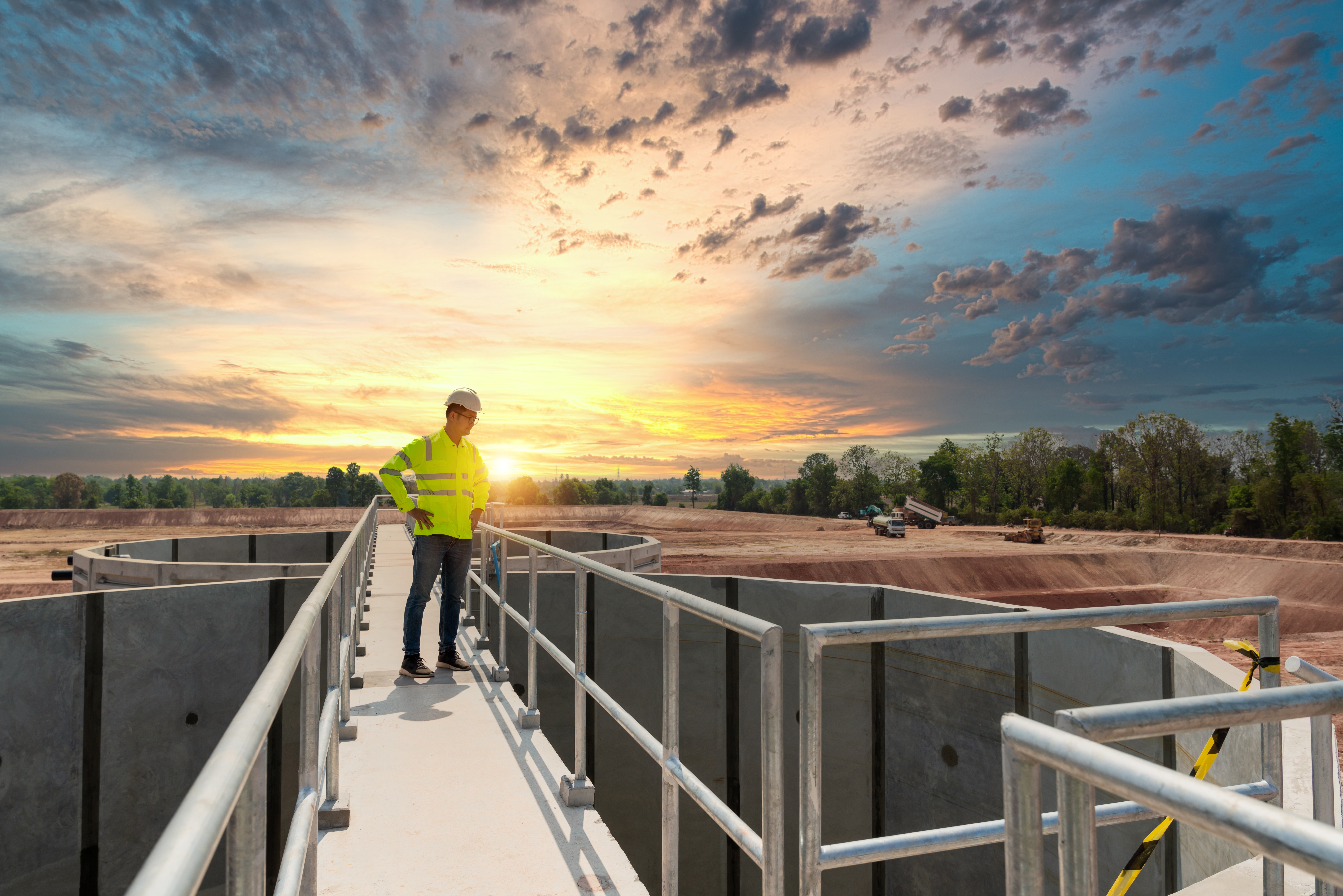 Wastewater treatment operator in a hard hat and safety jacket inspecting open treatment tanks at a facility